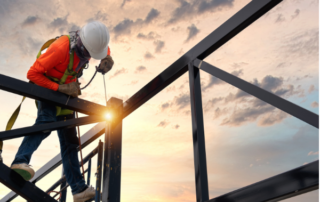 Untitled design 12 | McCormick Insurance Construction worker in an orange safety vest and white helmet welding steel beams at a partially built frame during sunset